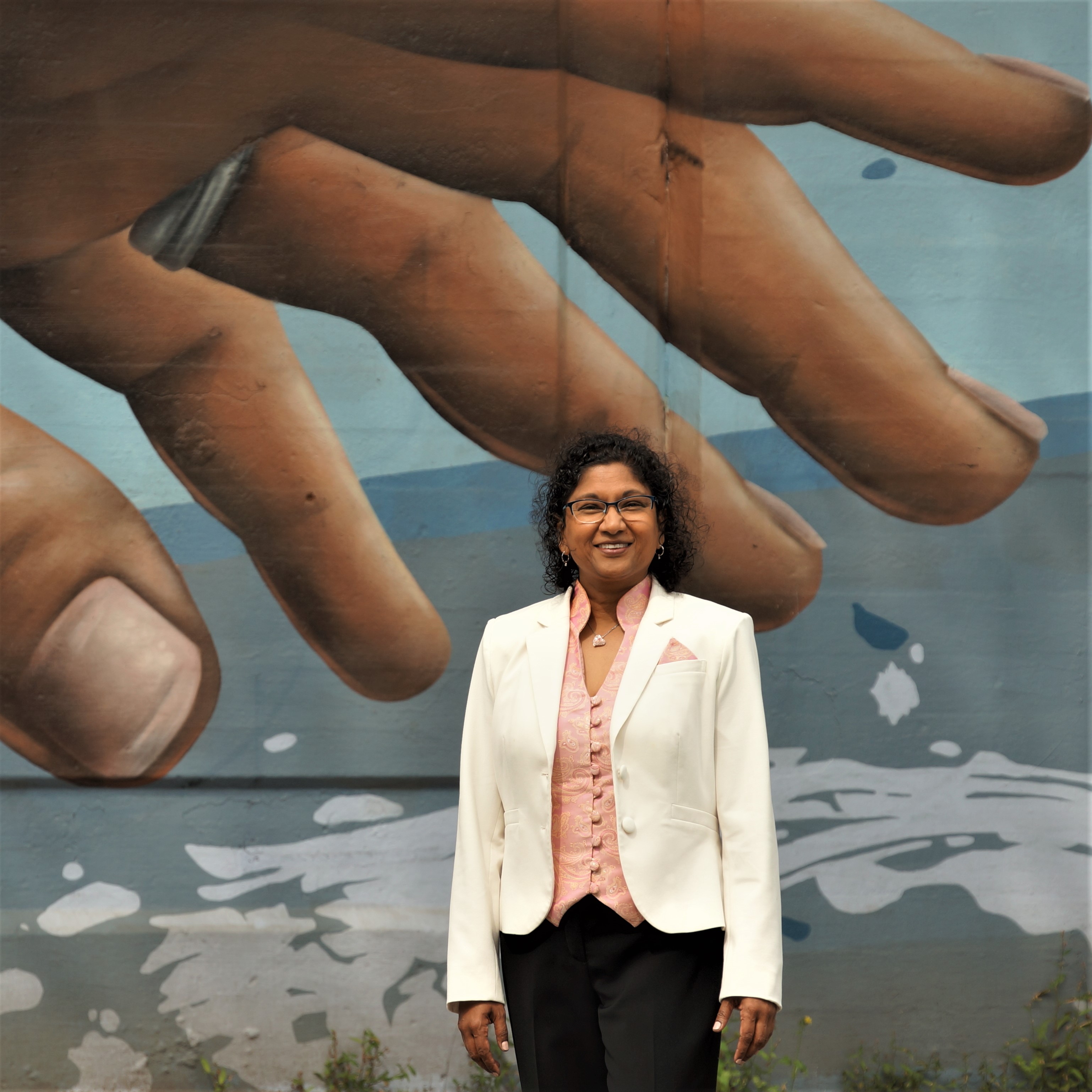 Wall mural of brown-skinned hand hovering over image of ME standing (ME = curly, black-haired, brown-skinned, smiling woman with glasses wearing a suit (white jacket, black pants and pink and gold paisley vest Wall mural of brown-skinned hand hovering over image of ME standing (ME = curly, black-haired, brown-skinned, smiling woman with glasses wearing a suit (white jacket, black pants and pink and gold paisley vest
